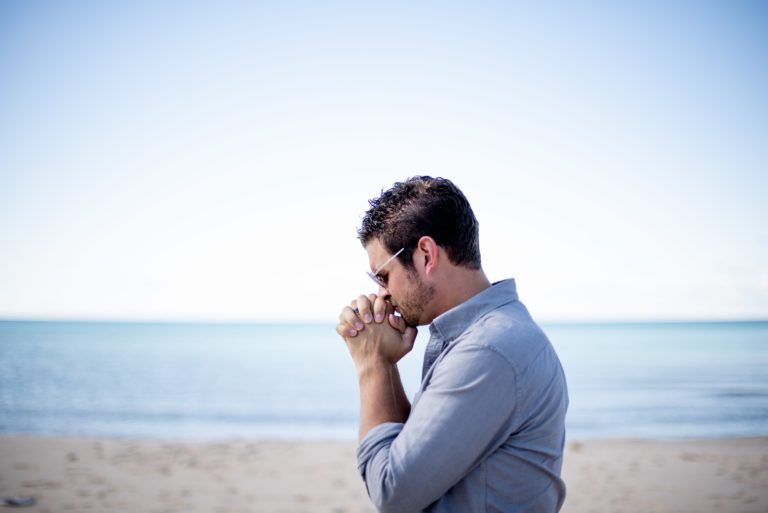 Man Kneeling Alone On A Beach With His Head Bowed In Prayer