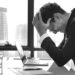 Man Sitting At His Desk Holding his Head In His Hands After Realizing That He Made A Huge Mistakes